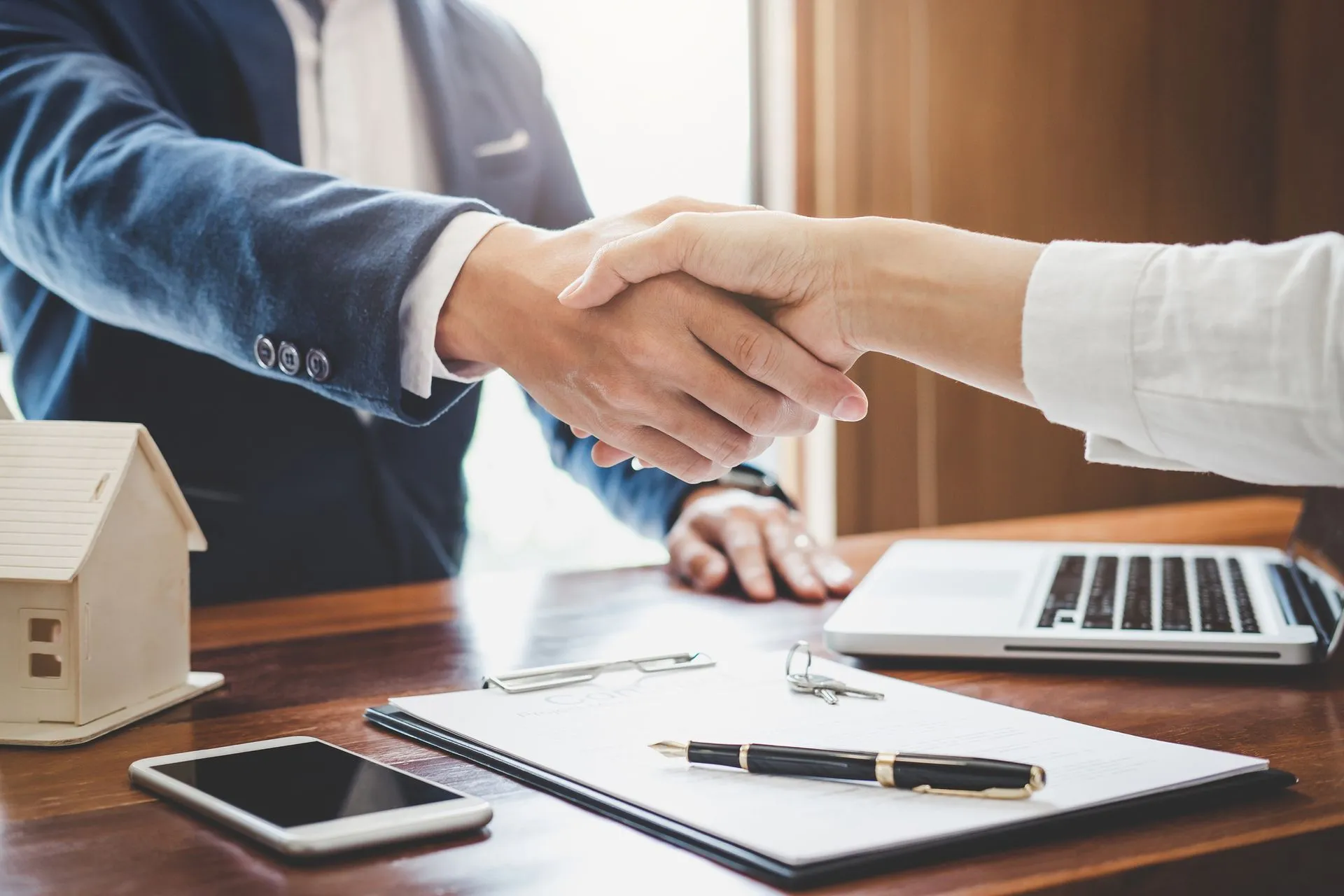 Two people shaking hands over a desk with a house model and paperwork.