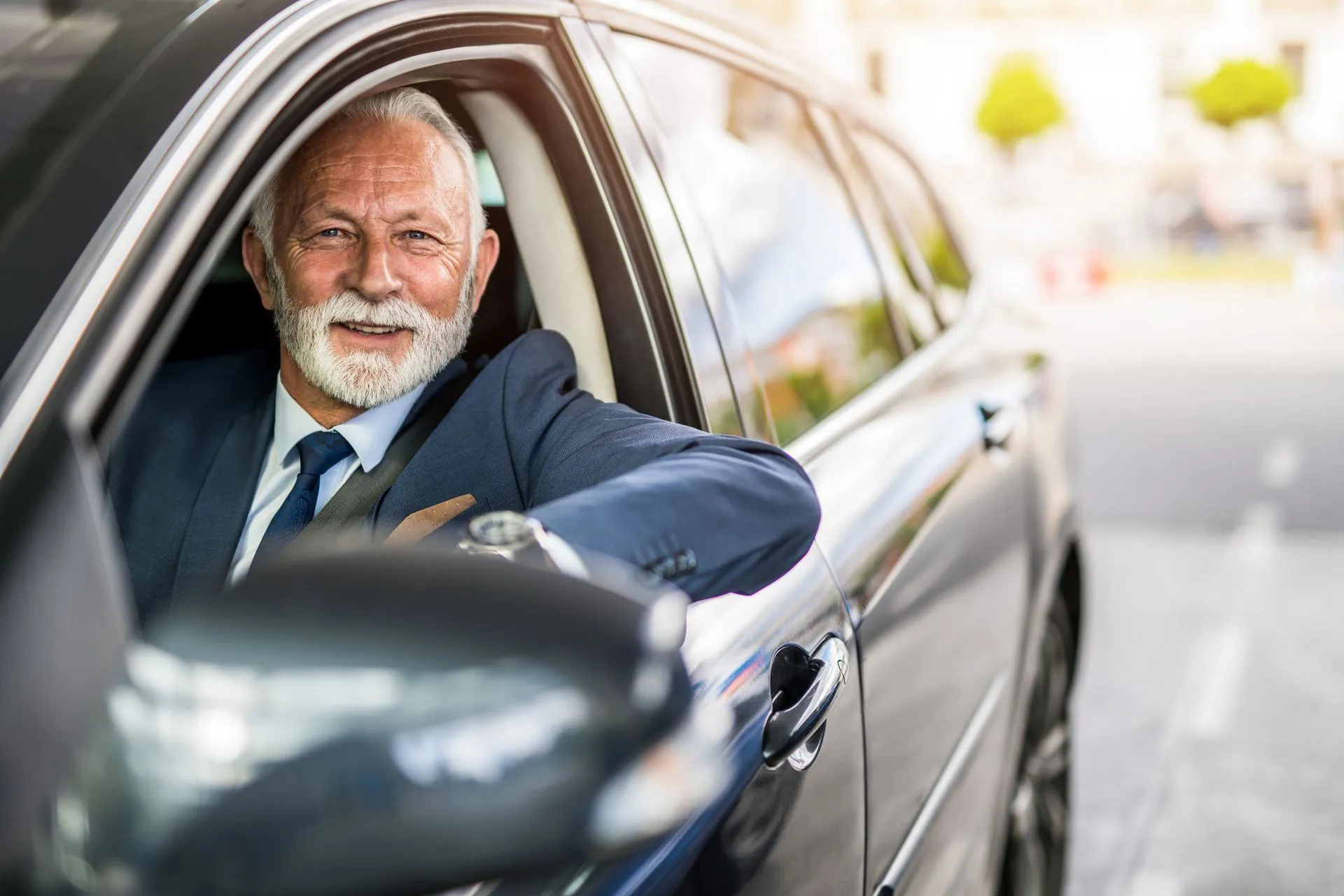Smiling older man in a suit driving a gray car.