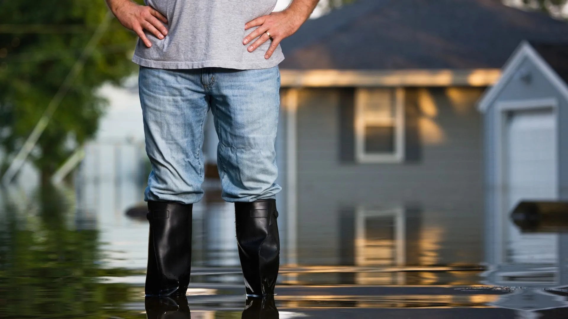 Man in boots standing in floodwater with a house in the background.