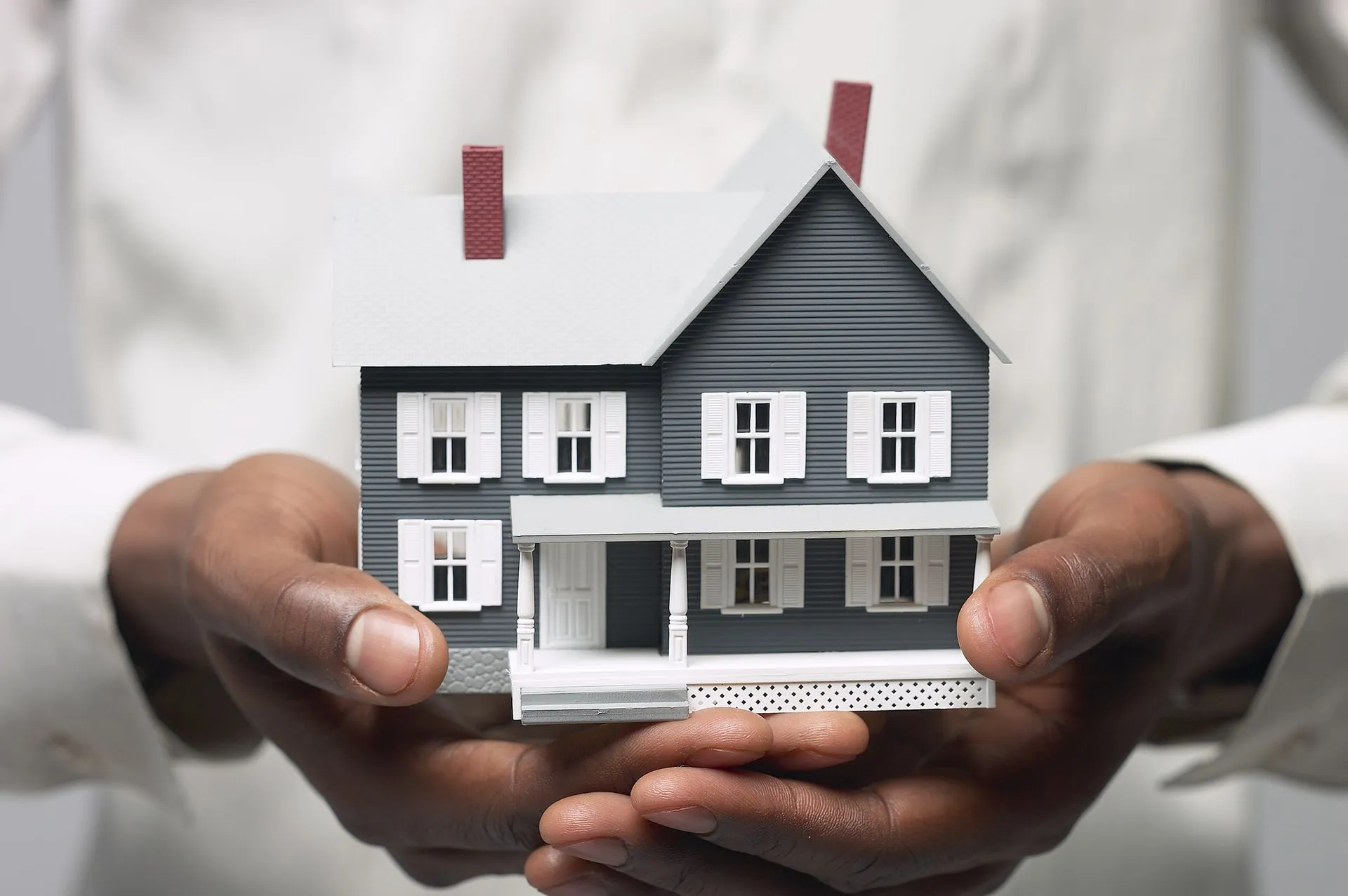 Person holding a model house with gray siding and a red chimney.