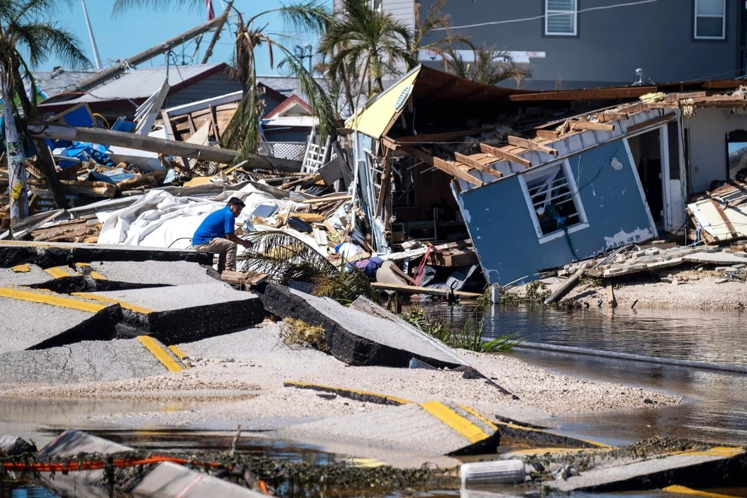 Man sitting amid rubble after a hurricane, with destroyed houses nearby.